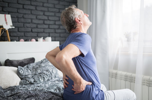 grey haired man sitting on bed holding lower back